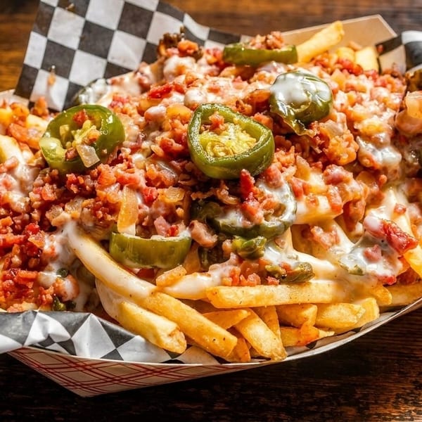 Loaded fries with melted cheese, bacon bits, and sliced jalapeños in a paper tray.