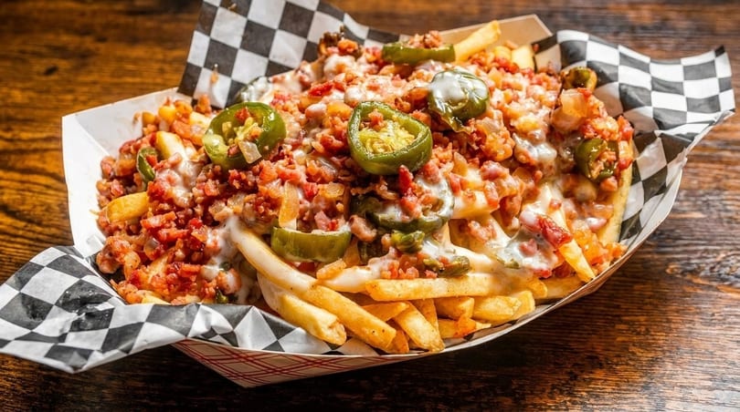 Loaded fries with melted cheese, bacon bits, and sliced jalapeños in a paper tray.