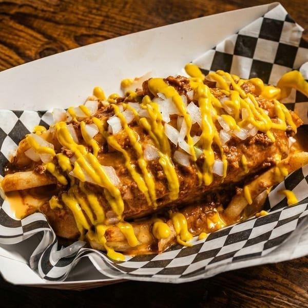 Chili cheese fries with diced onions and mustard in a checkered paper tray on a wooden table.