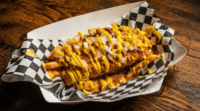 Chili cheese fries with diced onions and mustard in a checkered paper tray on a wooden table.
