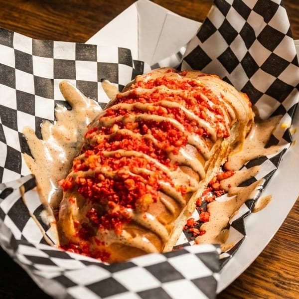 A hot dog topped with sauce and red crunchy bits, served in a checkered paper tray.