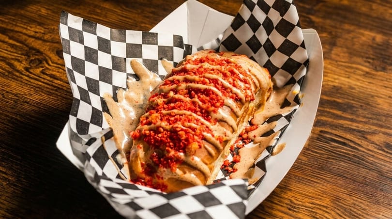 A hot dog topped with sauce and red crunchy bits, served in a checkered paper tray.